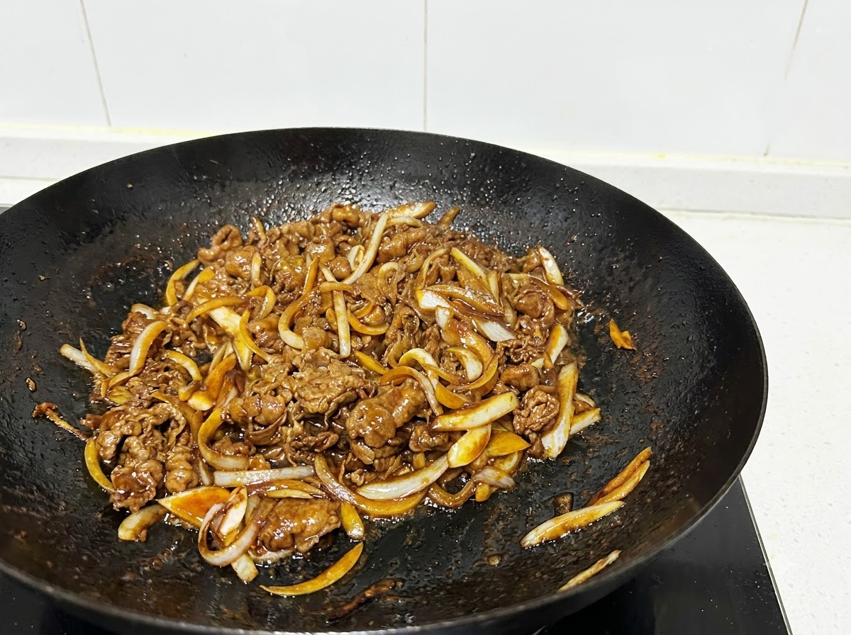 Primo piano di fettine di manzo e strisce di cipolla saltate in un wok nero con utensili da cucina.