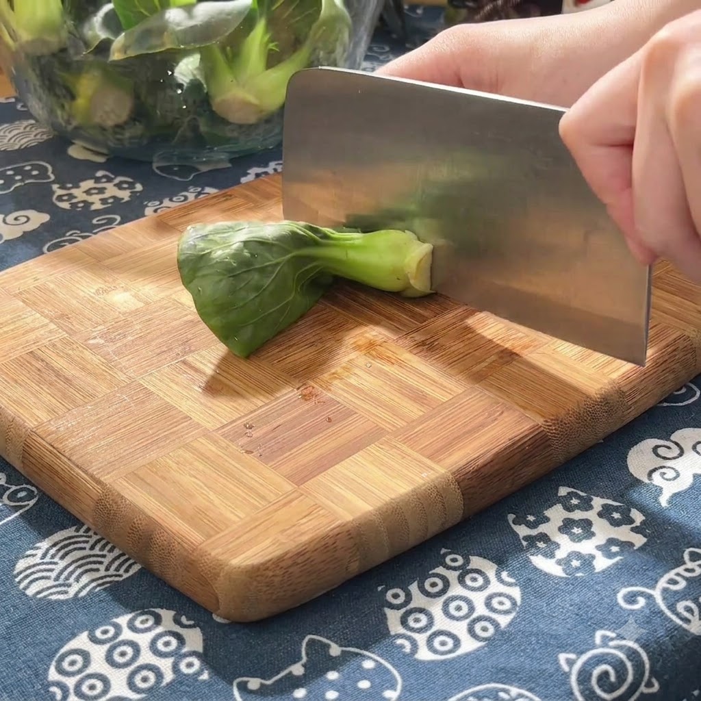Verdure tenere verdi tagliate a piccoli pezzi su un tagliere di legno usando una mannaia pesante.