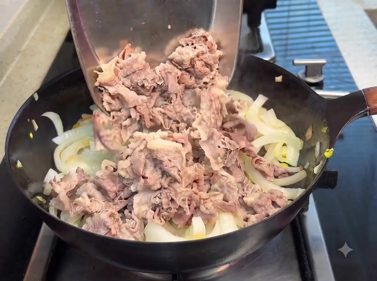 Fette di manzo sbollentate versate da una spatola di metallo in una padella di cipolle traslucide, con salsa teriyaki versata sopra.