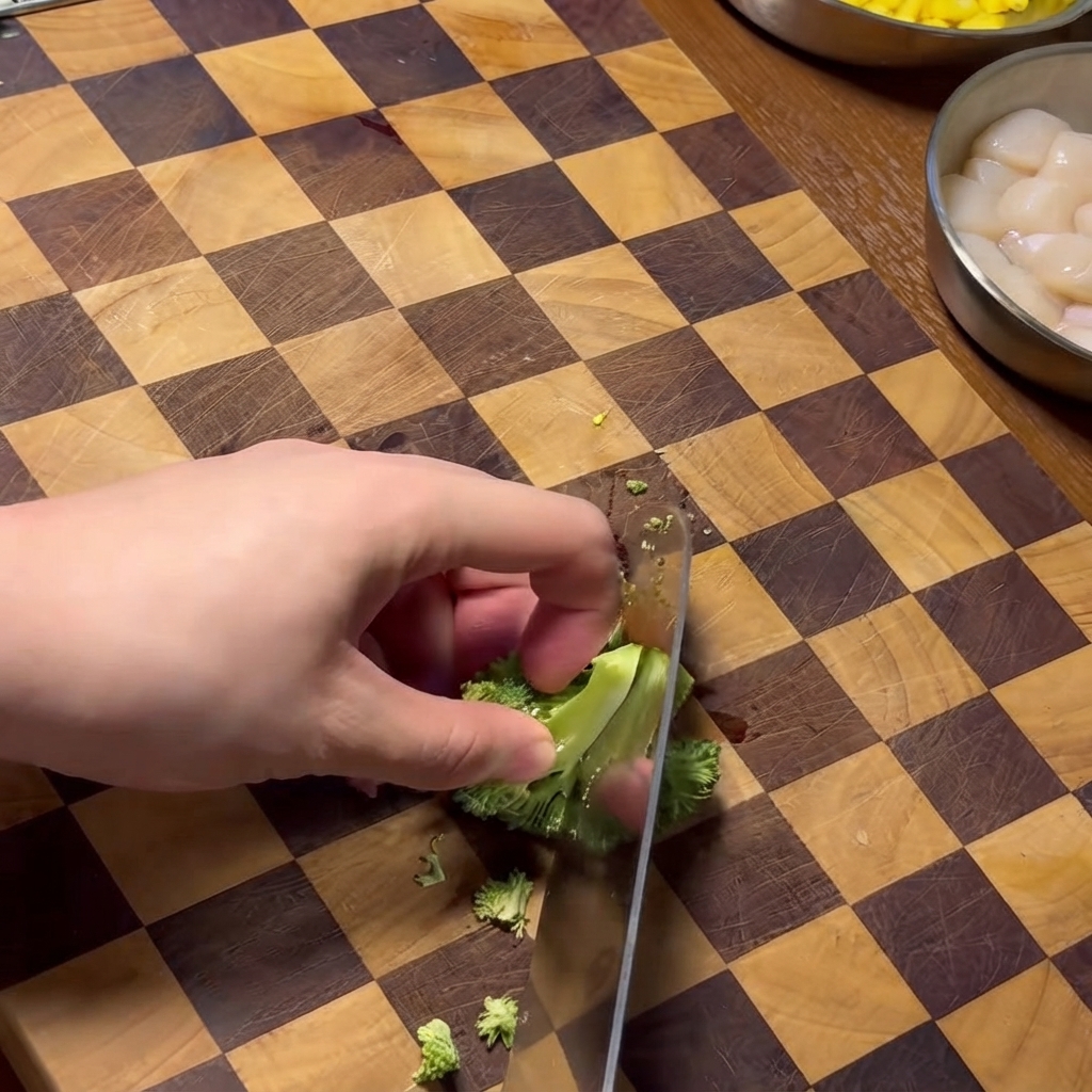 Una mano taglia al coltello le cimette di broccoli su un tagliere in legno a scacchi.
