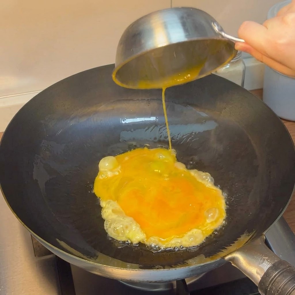 Uova sbattute versate da una ciotola dacciaio in un wok rovente.