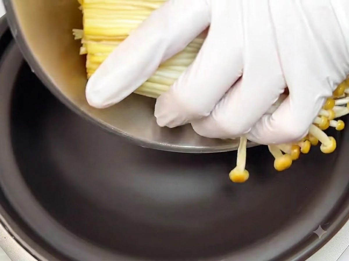 Mani con i guanti che posizionano un mazzetto di funghi enoki crudi mondati sul fondo di un tegame di coccio nero.