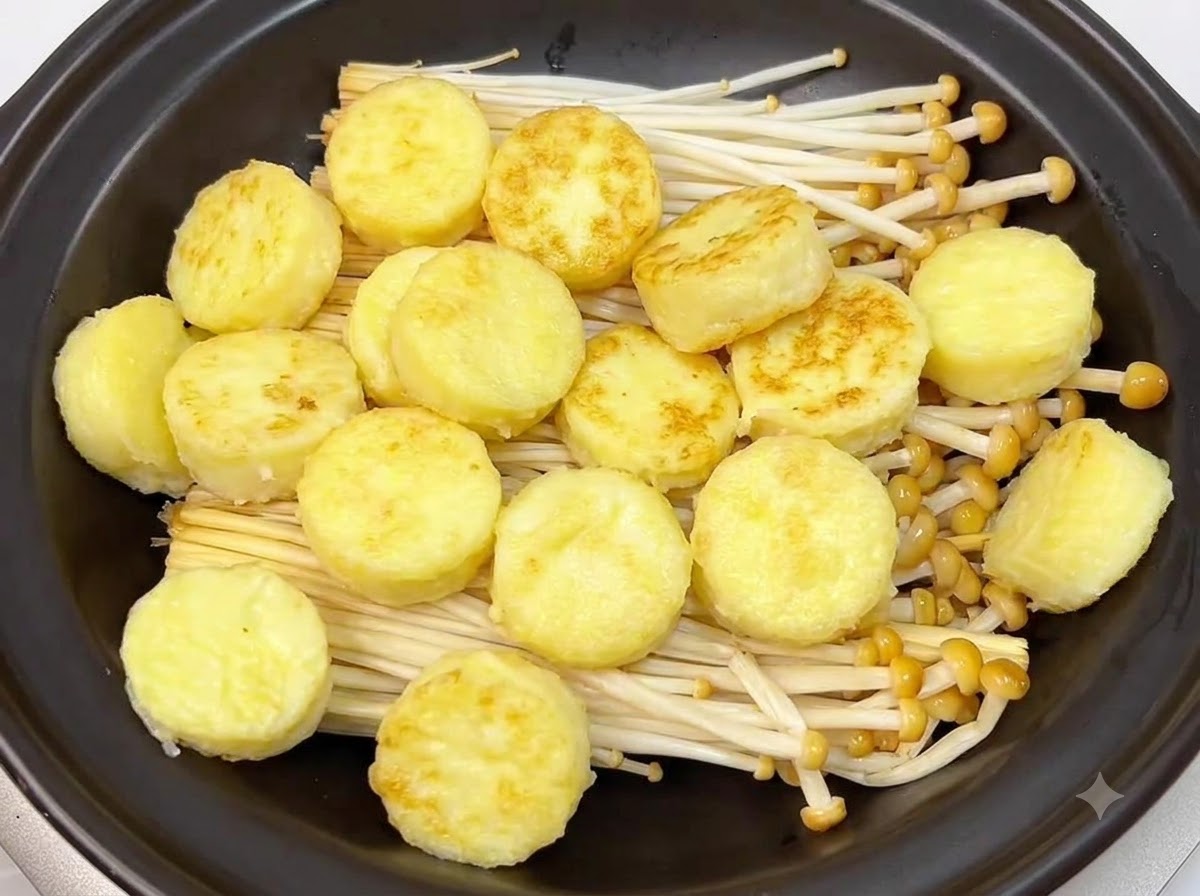 Rondelle dorate di tofu giapponese fritte disposte ordinatamente sopra i funghi enoki allinterno di un tegame di coccio nero.