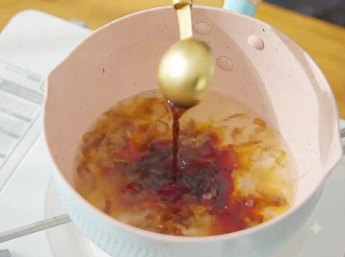 Versare la salsa di soia scura da un cucchiaio dorato nel brodo di bonito in ebollizione nella pentola rosa.