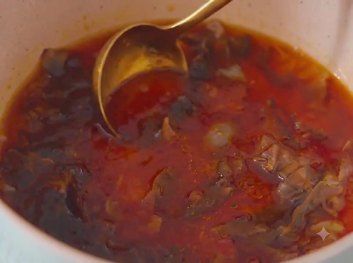 Un primo piano di un cucchiaio di metallo dorato che mescola una miscela di salsa marrone rossastra con fiocchi di bonito allinterno di una pentola di colore chiaro.