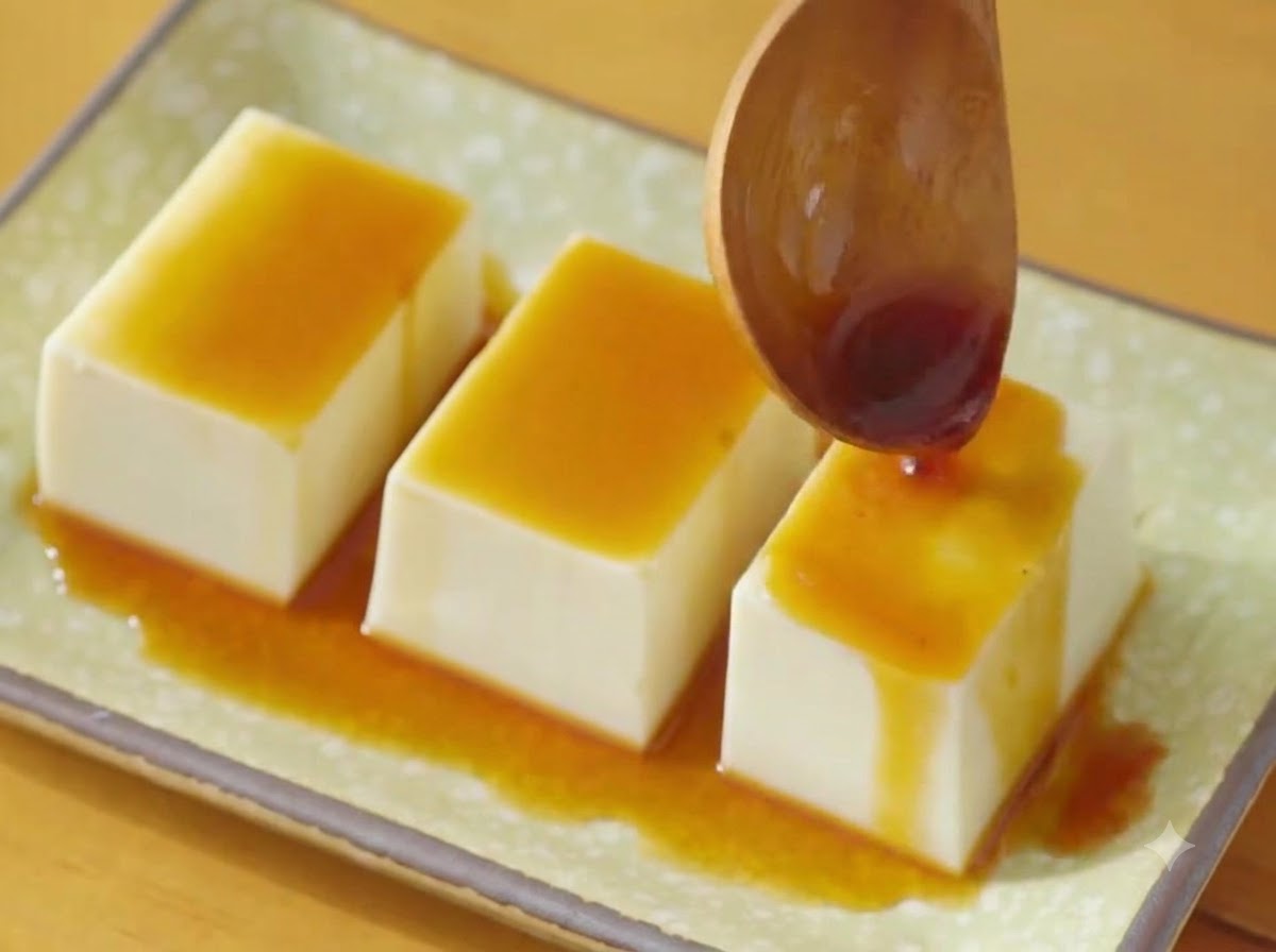 Un cucchiaio di legno che versa salsa di soia scura su tre file di tofu silken su un piatto verde.