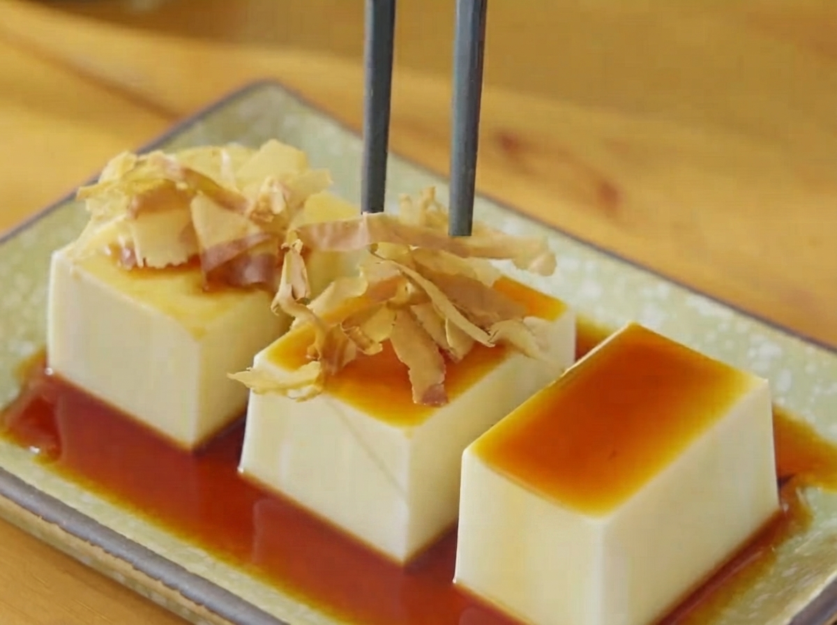 Bacchette che mettono fiocchi di bonito secchi e strisce di alghe sul tofu condito con soia.
