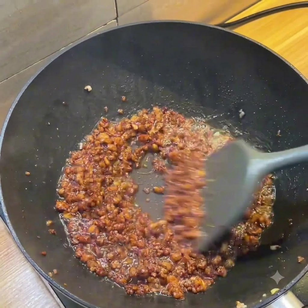 Un wok scuro che mostra una ricca salsa di carne macinata marrone mescolata con una spatola grigia.