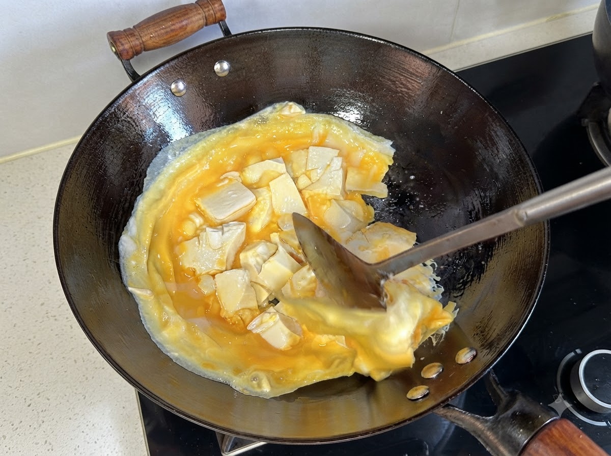 Una spatola che solleva e capovolge lo strato cotto di uova e tofu nel wok.