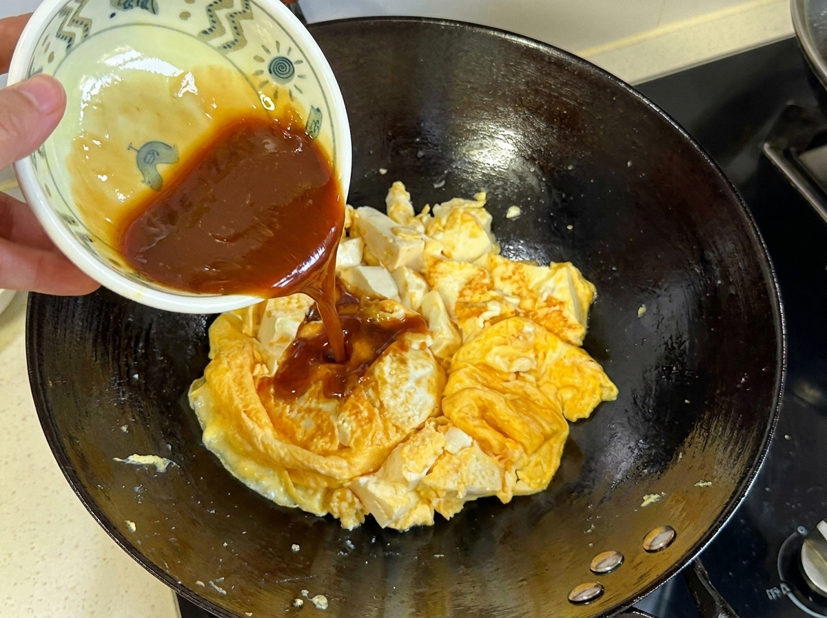 Salsa marrone scuro versata da una ciotolina sul composto cotto di uova e tofu nel wok.