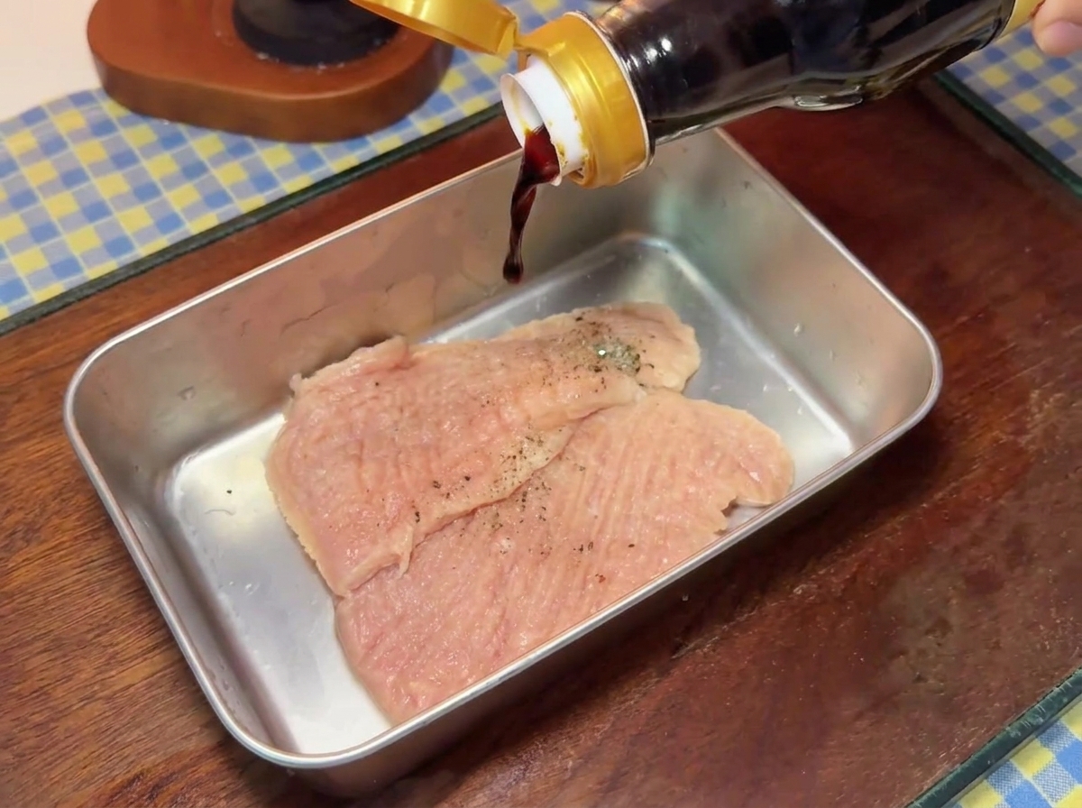 Salsa di soia scura che cola da una bottiglia di vetro direttamente sulle cotolette di maiale condite dentro un vassoio di metallo.