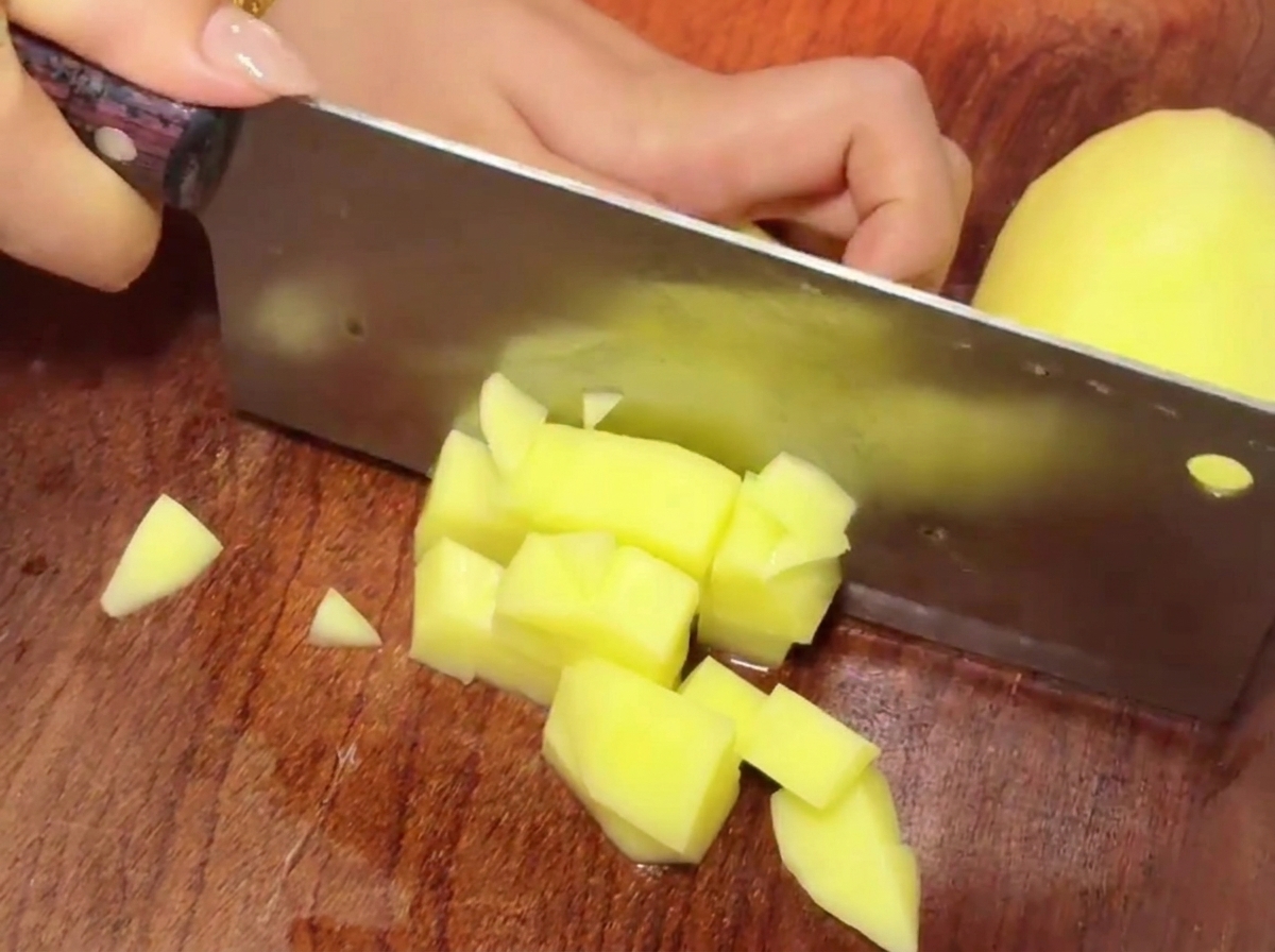 Una mannaia che taglia pezzi di patate gialle su un tagliere di legno per creare piccoli cubi.