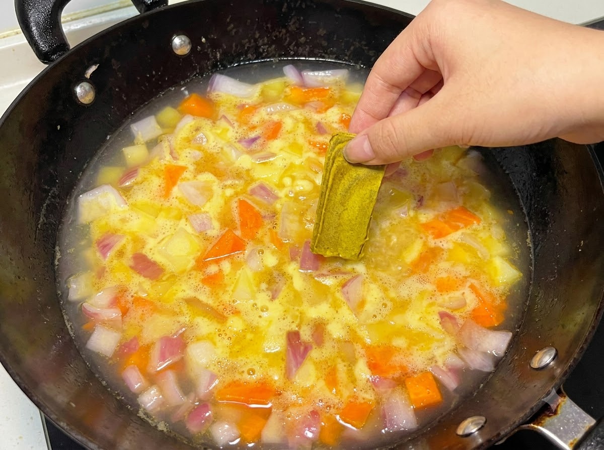 Una mano che posiziona un blocchetto solido di curry in una pentola di acqua e verdure che sobbollono.