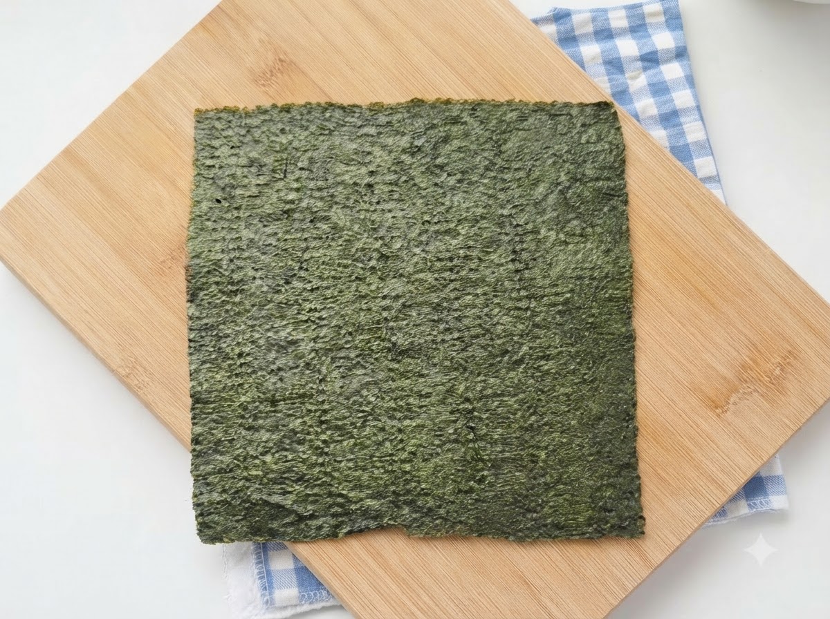 Un foglio intero di alga nori da sushi appoggiato su un tagliere di legno con un panno a quadretti blu sotto.