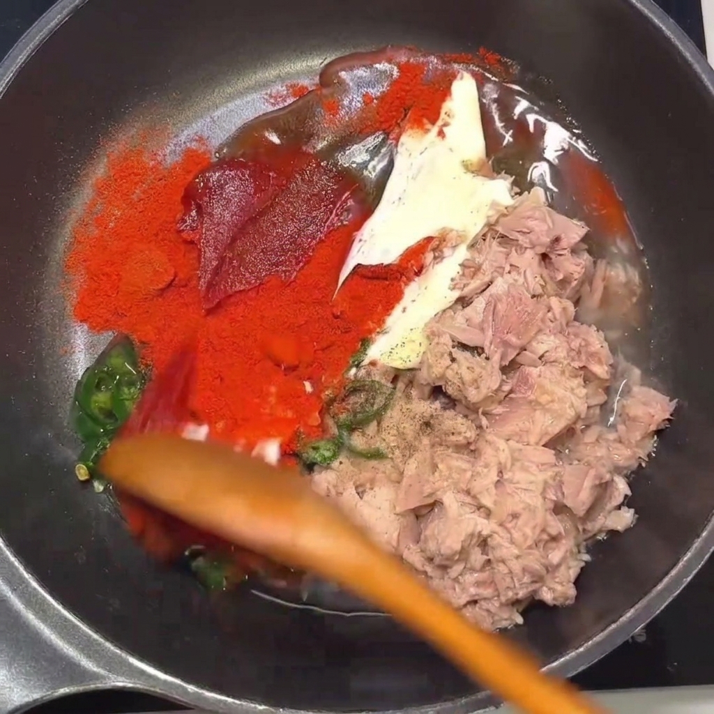 Un cucchiaio di legno che mescola il tonno, il peperoncino e i condimenti in una padella.