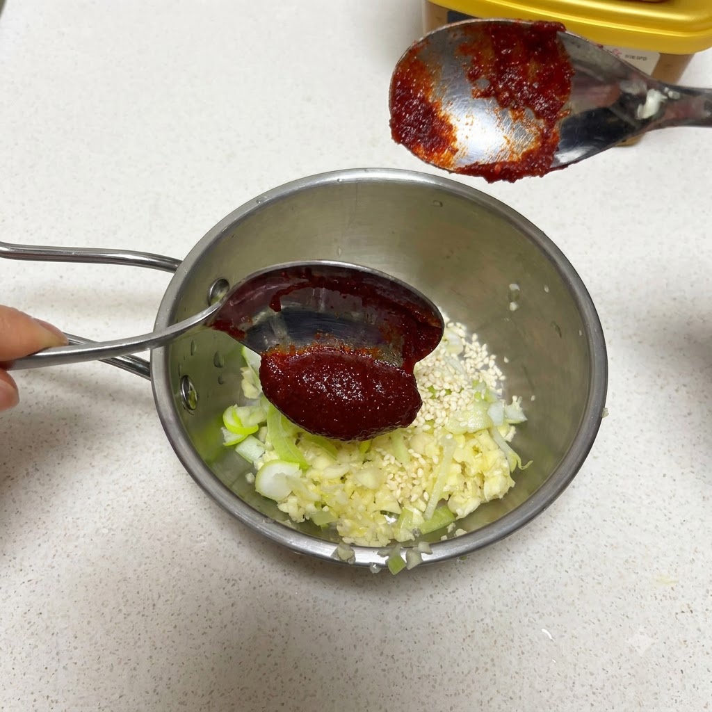 Un cucchiaio di pasta di peperoncino coreana tenuto sopra una padella con aglio e semi di sesamo.