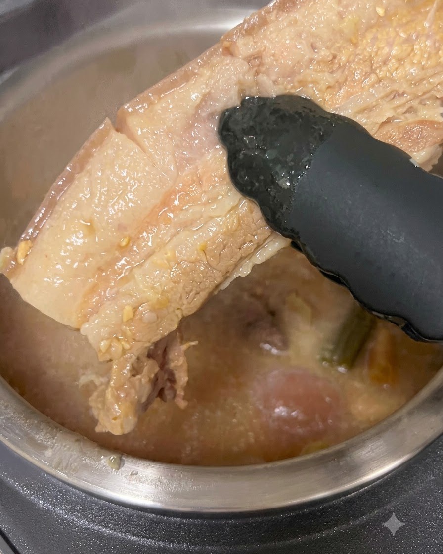 Un pezzo spesso di pancetta di maiale cotta estratto da una pentola di brodo con pinze nere.
