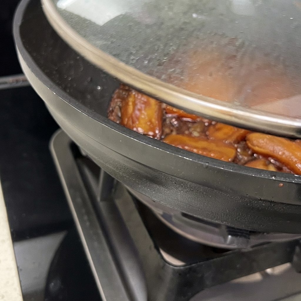 Primo piano di un wok con il coperchio parzialmente sollevato, che mostra le costine di maiale che cuociono in una salsa bruna densa e lucida.