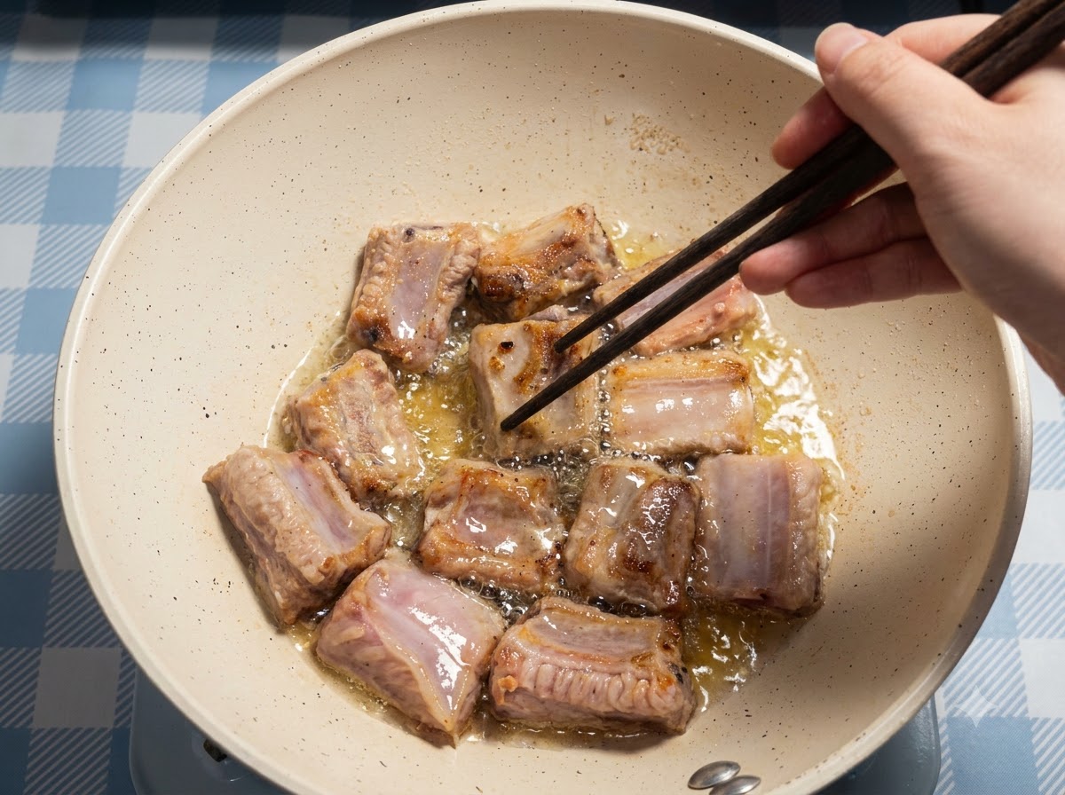 Molteplici pezzi di costine di maiale che friggono nellolio dentro una padella, con una mano che usa le bacchette per sistemarle.
