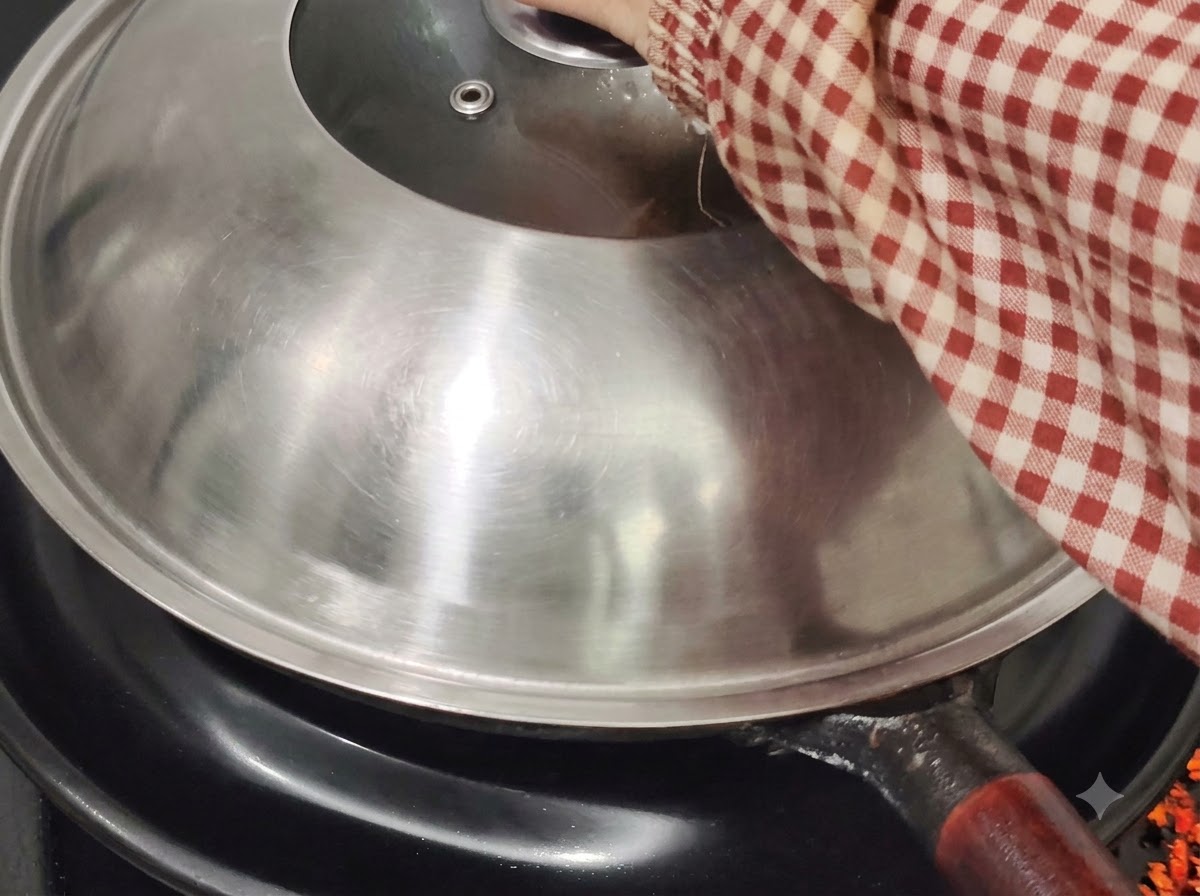 Un coperchio di metallo per wok che copre il wok su un piano cottura, tenuto da una mano con una manica a quadri rossi e bianchi.