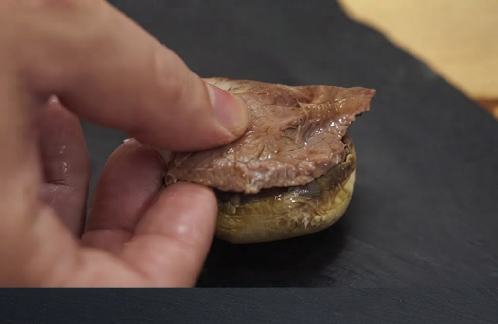 Una mano che posiziona un pezzo di carne cotta su una base di cappella di fungo cotta.