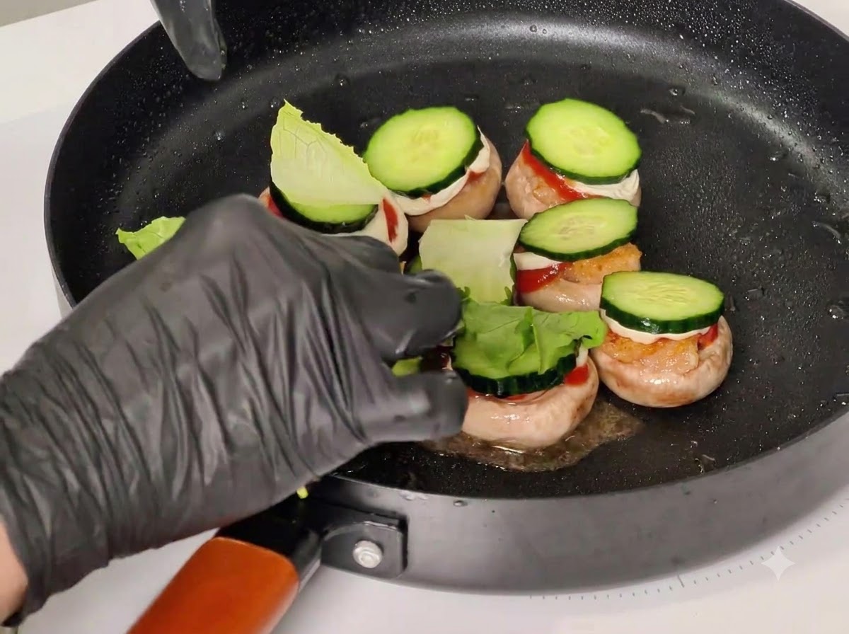 Una mano guantata che inserisce stuzzicadenti attraverso gli ingredienti impilati nelle cappelle dei funghi allinterno di una padella nera.