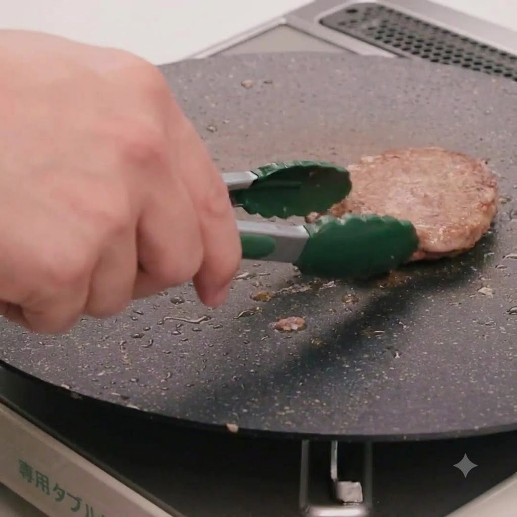 Una mano che utilizza pinze da cucina verdi per girare un patty di manzo in cottura in una padella piatta.