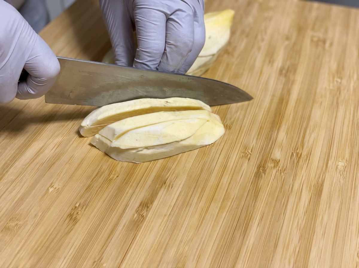 Patate dolci crude pelate tagliate a bastoncini lunghi con un coltello su un tagliere di legno.
