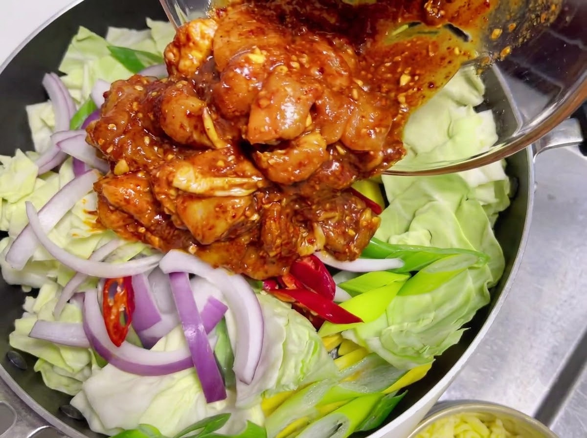 Pollo crudo marinato ricoperto di salsa rossa piccante versato da una ciotola di vetro su uno strato di cavolo e cipolle crudi.