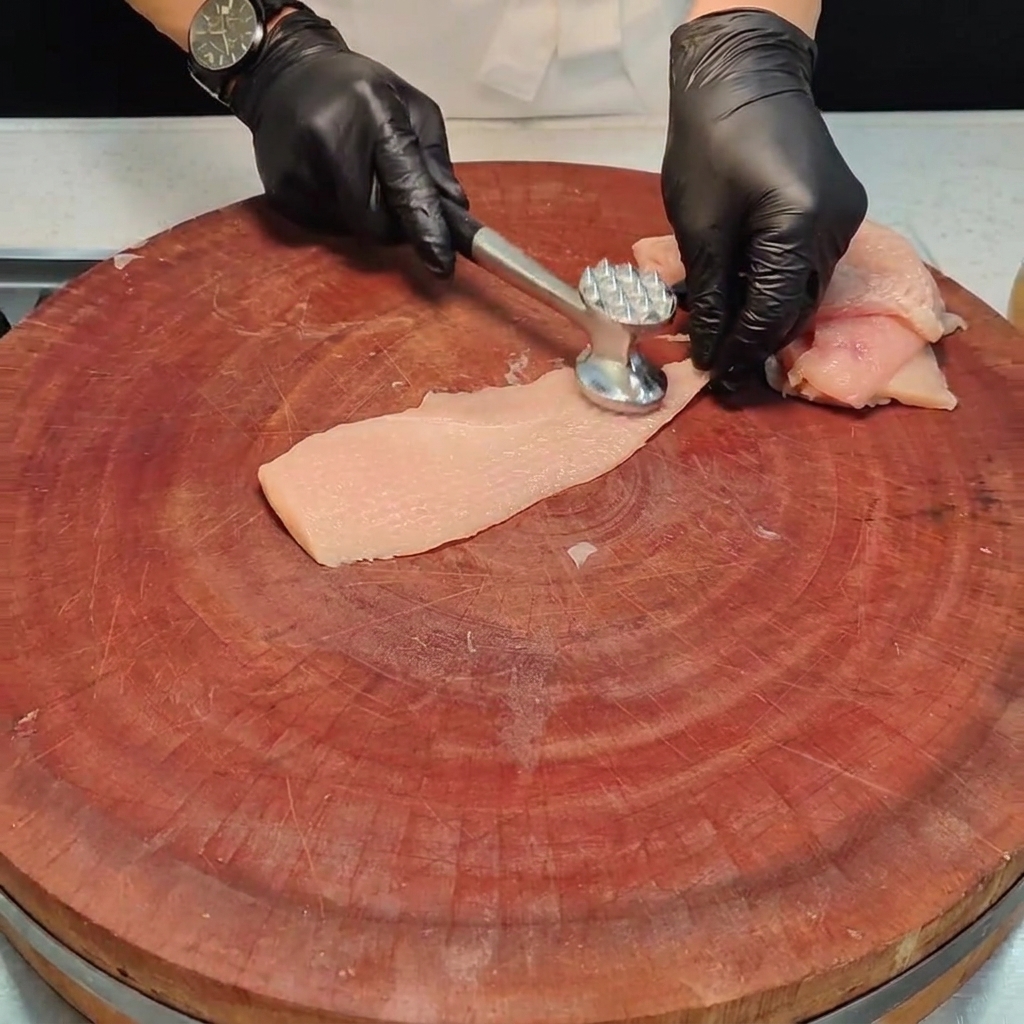 Un pezzo di petto di pollo battuto con un battere per carne in metallo su un tagliere di legno.