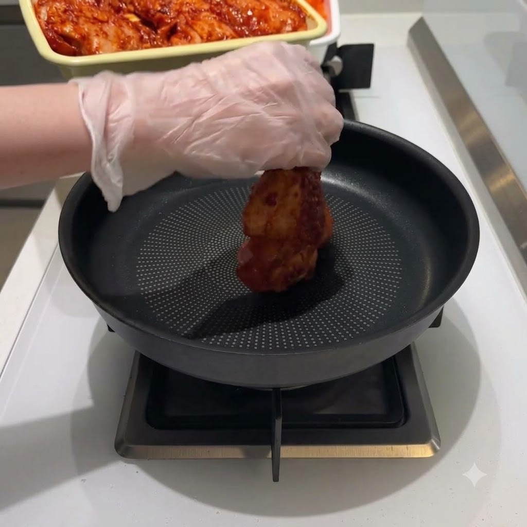 Una mano guantata che posiziona un pezzo di pollo marinato rosso al centro di una padella nera antiaderente posta sul fuoco.