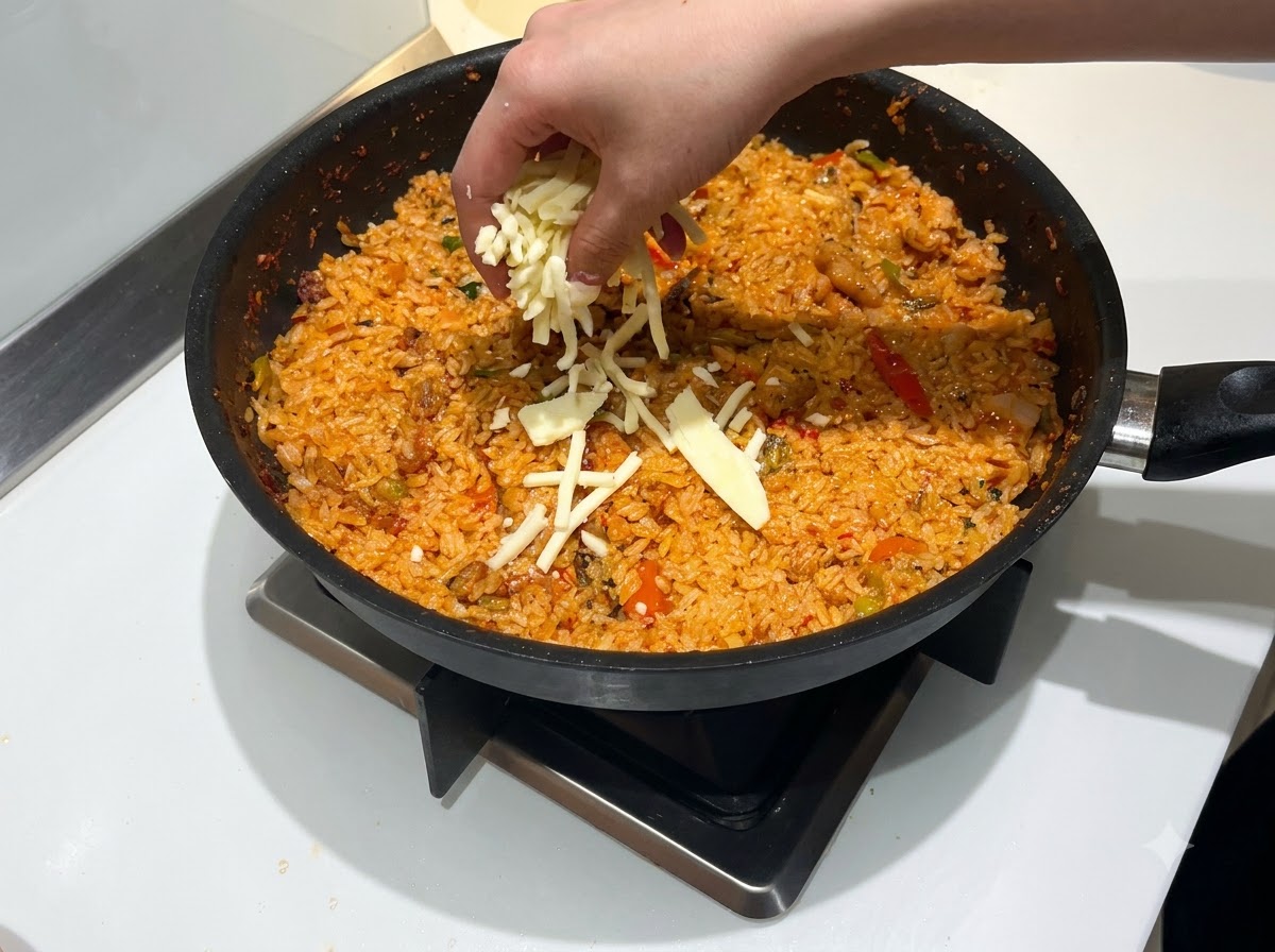 Una mano che sparge una generosa quantità di mozzarella grattugiata sopra una padella di riso saltato rosso e piccante.