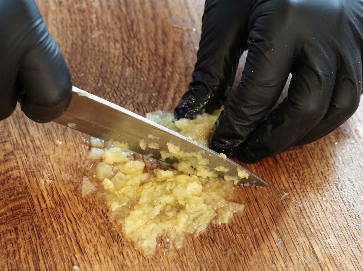 Un paio di mani guantate che usano un coltello per tagliare e ridurre in purea la polpa di limone avanzata su un tagliere di legno.