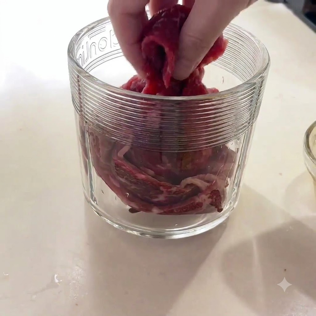 Una mano inserisce pezzi di carne cruda nel contenitore in vetro di un robot da cucina.