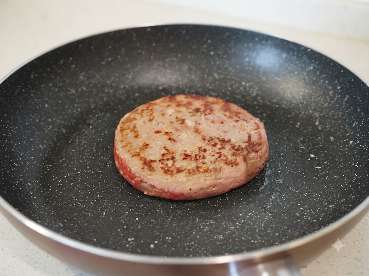 Un piccolo getto dacqua che viene versato in una padella accanto a una polpetta di hamburger parzialmente rosolata.