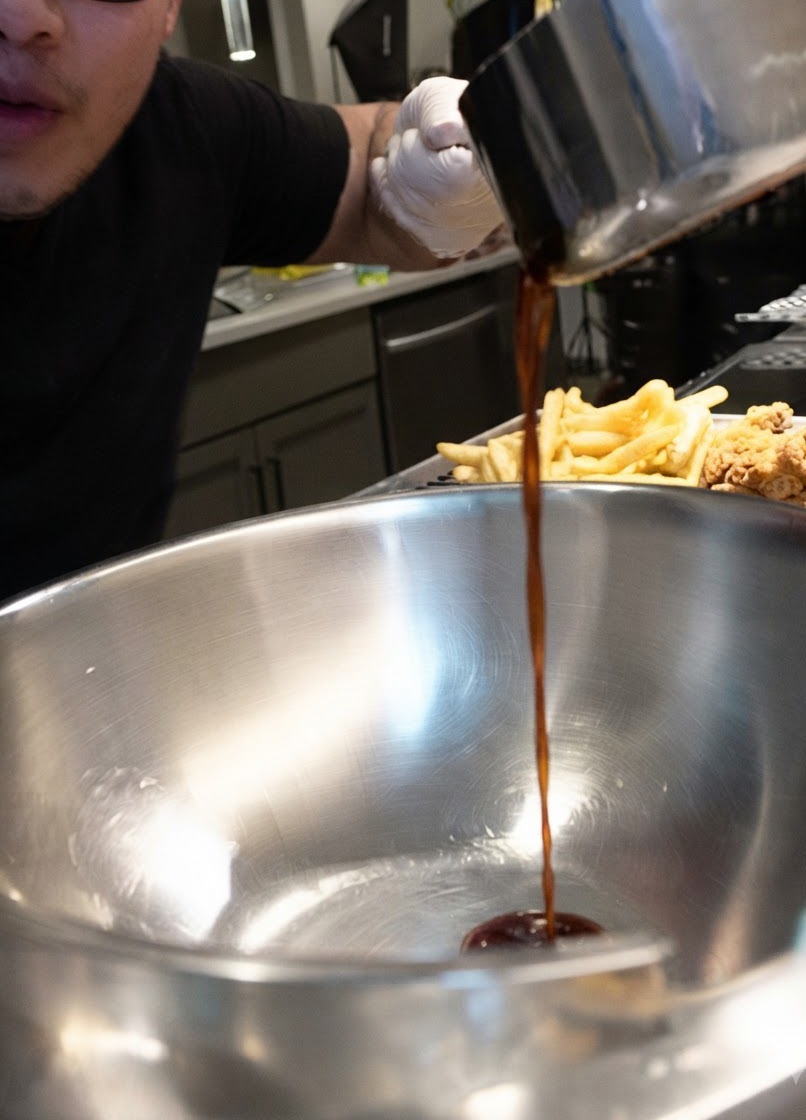 Salsa scura versata in un flusso costante da un pentolino in una grande ciotola dacciaio accanto a pezzi di pollo fritto.