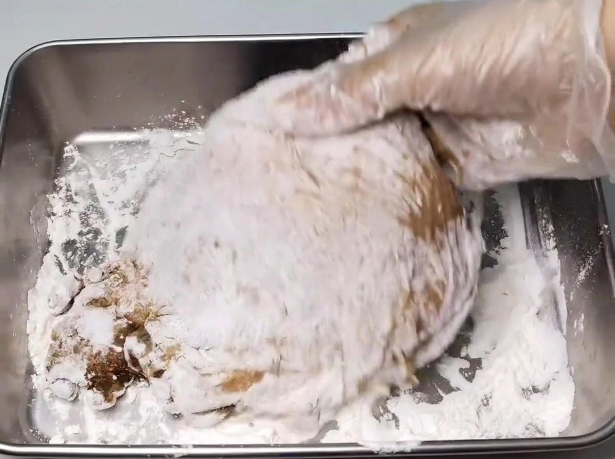 Una mano con un guanto che ricopre una coscia di pollo marinata con uno strato di amido bianco allinterno di un vassoio di metallo.