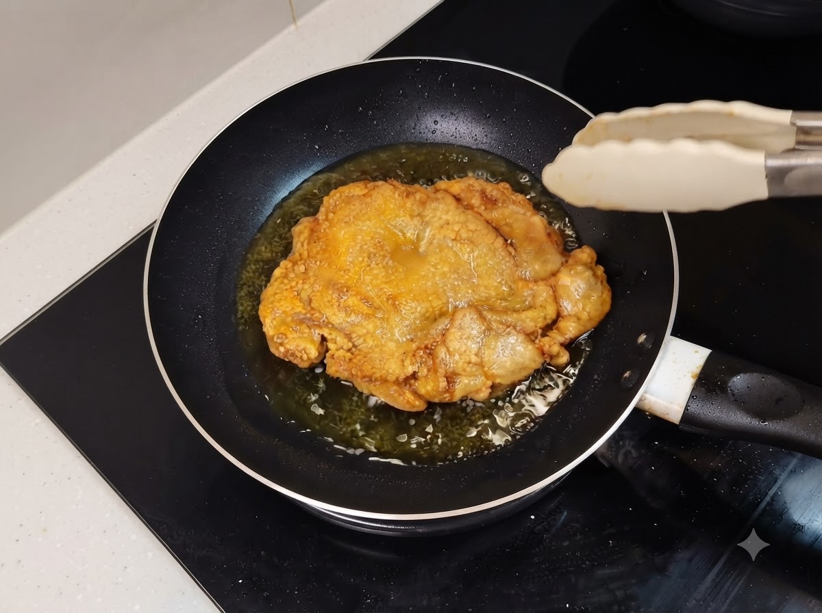 Una coscia di pollo fritta dorata che sfrigola in una padella oleosa con pinze da cucina vicine.