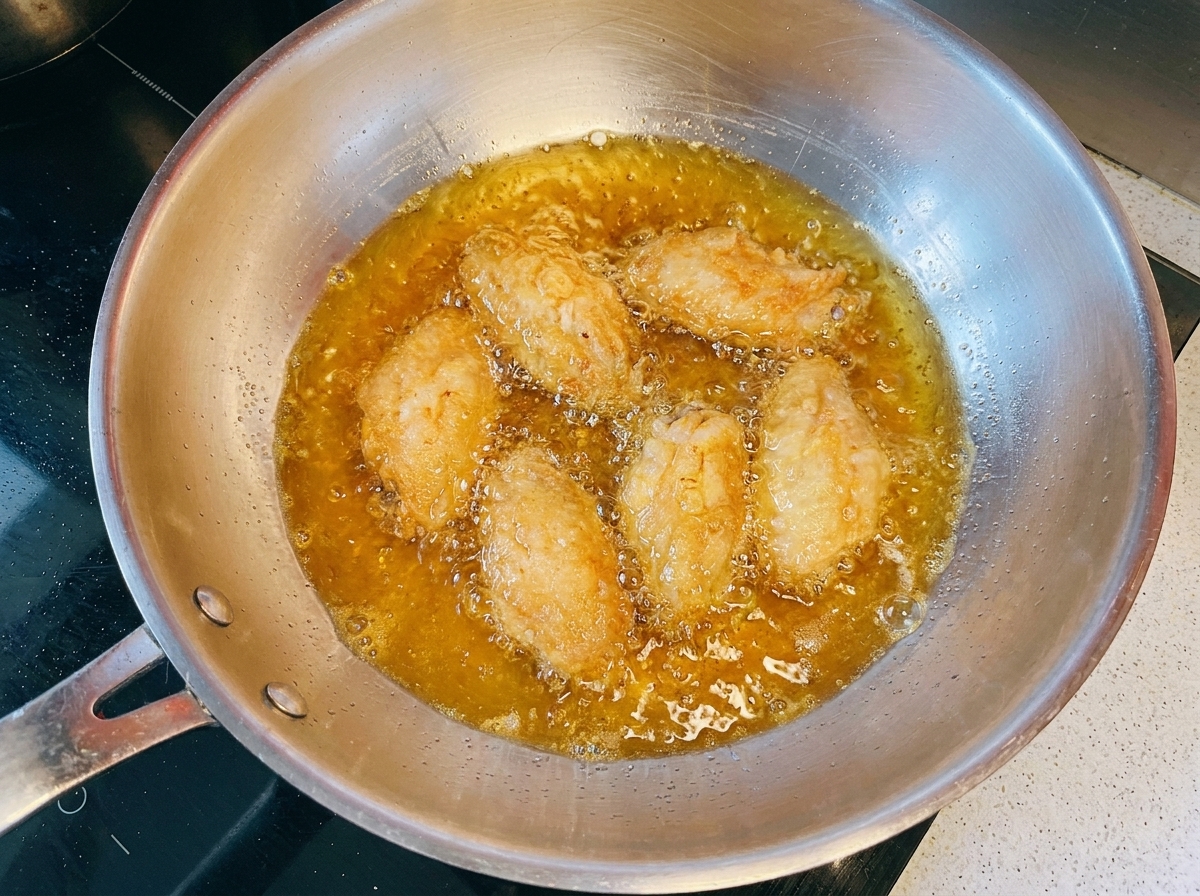 Ali di pollo dorate che friggono nellolio caldo allinterno di una padella di metallo.