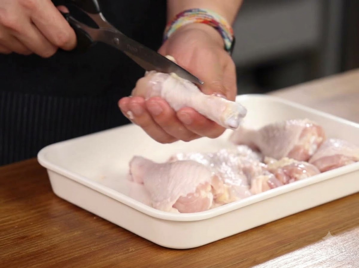 Mani che usano le forbici da cucina per tagliare attorno allosso di una coscia di pollo allinterno di un vassoio bianco.