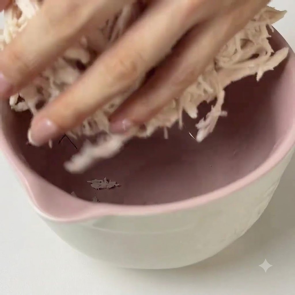 Mani che mescolano la salsa pesto verde nel pollo sfilacciato dentro una ciotola rosa.