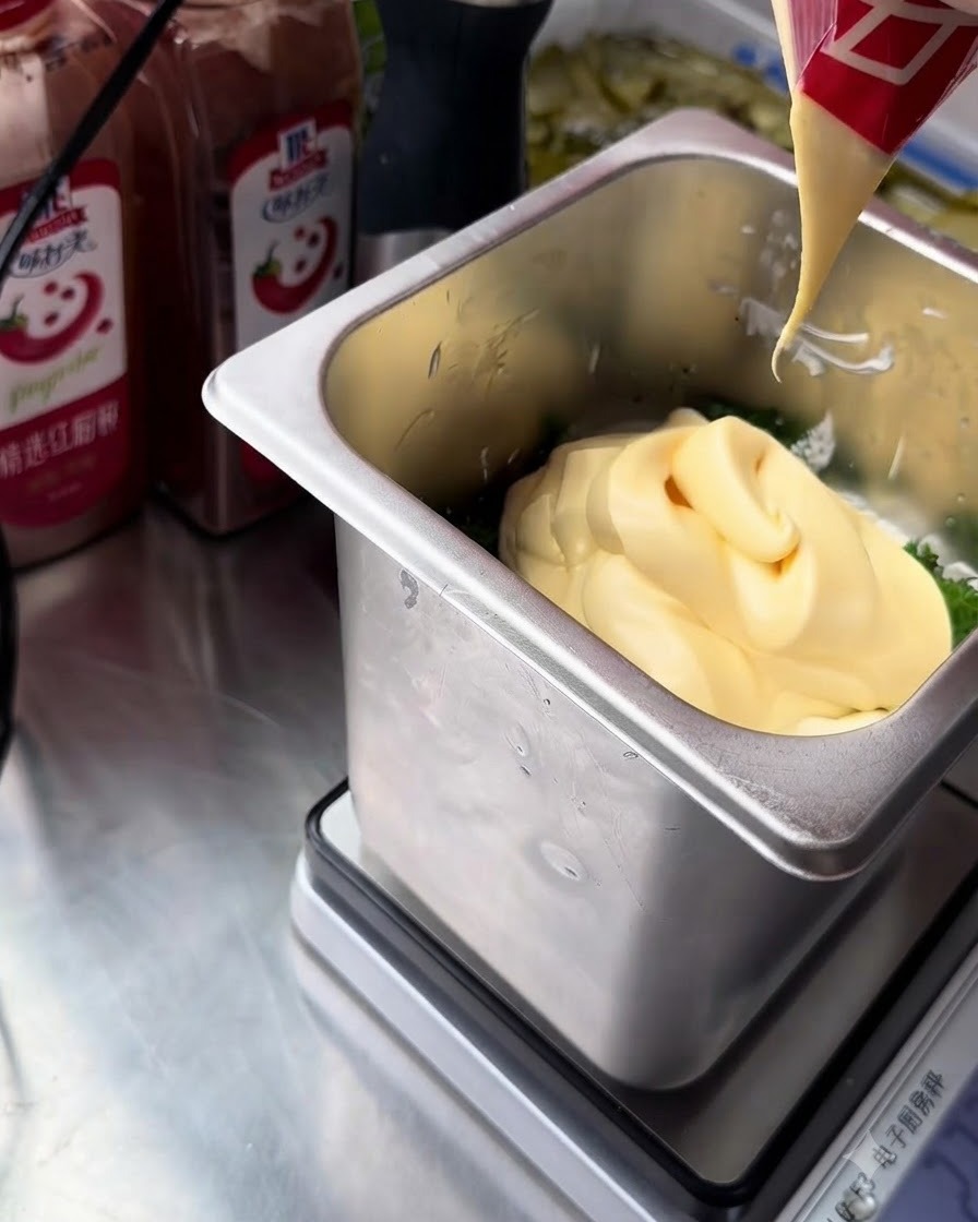 Un flusso di maionese ricca con tuorlo duovo giallo spremuto da una confezione rossa e bianca nel contenitore in acciaio inossidabile con gli ingredienti della salsa tartara.